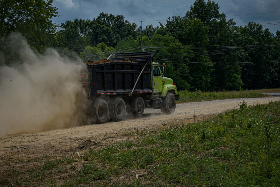 Dump truck carrying soil driving along an unpaved construction road, kicking up a large cloud of dust with trees and utility lines in the background.
